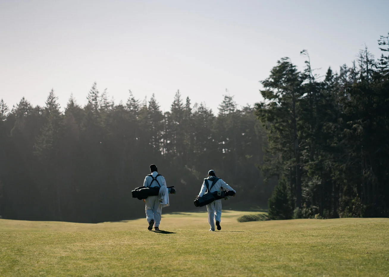 Two golf caddies walking away on a grassy golf course with a forest in the background.