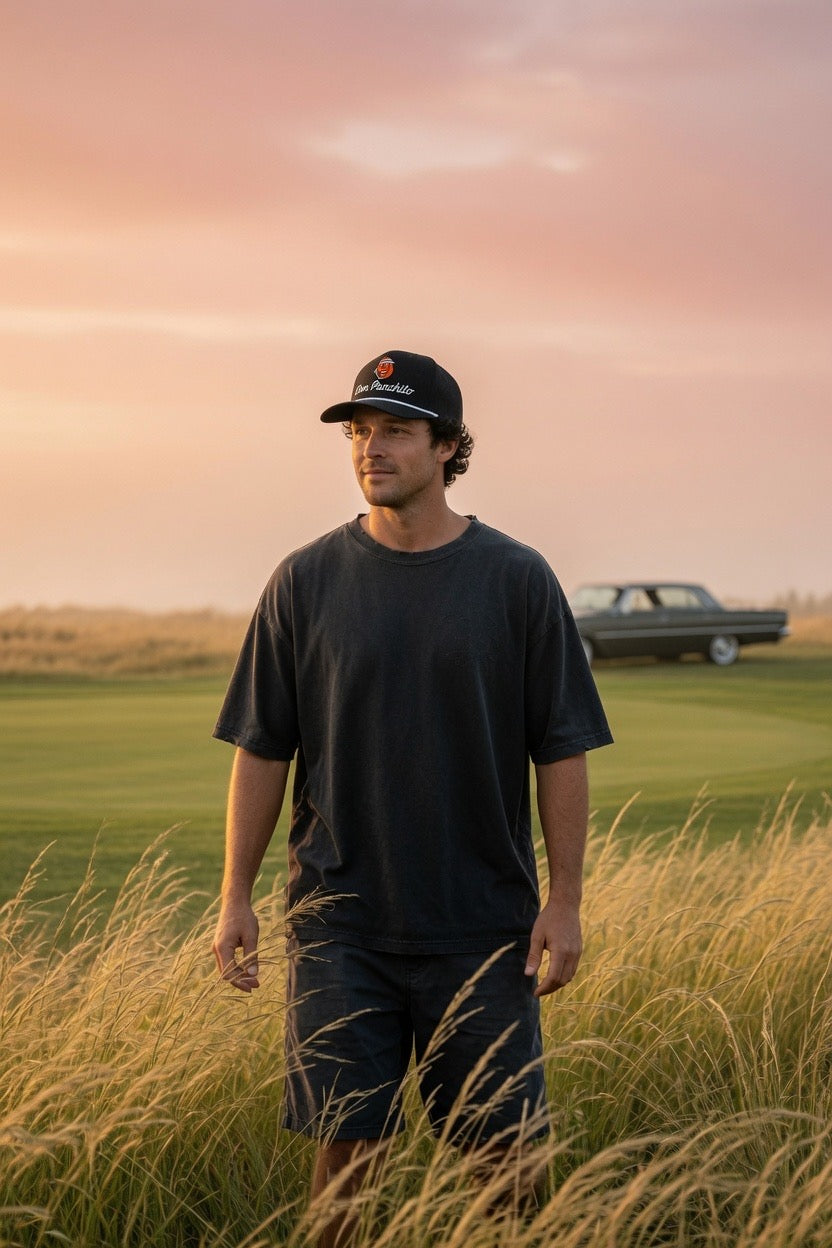 A man wearing a black t-shirt and black shorts stands in a field of tall dry grass at sunset, wearing a black DON PANCHITO rope hat. In the background, there is a golf course and a vintage car under a pink sky.