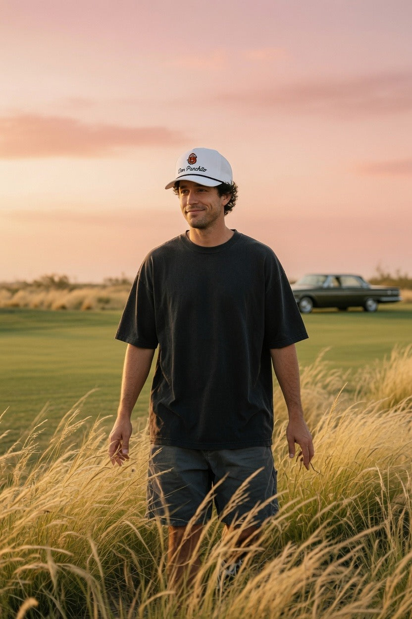 A man wearing a white baseball cap with a DON PANCHITO logo, a black t-shirt, and dark shorts stands in a field of tall dry grass at sunset, with a golf course and a vintage car in the background.