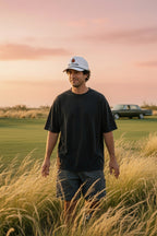 A man wearing a white baseball cap with a DON PANCHITO logo, a black t-shirt, and dark shorts stands in a field of tall dry grass at sunset, with a golf course and a vintage car in the background.