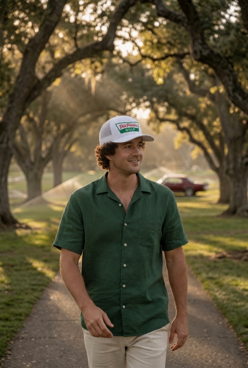 A man walking on a tree-lined path wearing a green short-sleeve button-up shirt, beige pants, and the white DON PANCHITO KRISPY trucker hat.
