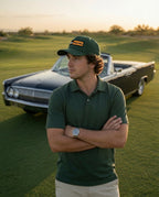 A man stands on a golf course wearing a dark green polo shirt, the matching green DON PANCHITO LATENIGHT dad hat, and light khaki pants, with a vintage black convertible car behind him at sunset.