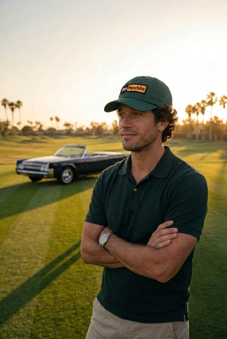 A man wearing the green DON PANCHITO LATENIGHT dad hat and a dark green polo shirt stands on a golf course with his arms crossed, with a vintage convertible car in the background at sunset.