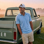 A man wearing a light blue polo shirt, khaki shorts, and the white DON PANCHITO NEON RACER trucker hat leans against the back of a vintage green pickup truck in a grassy field at sunset.