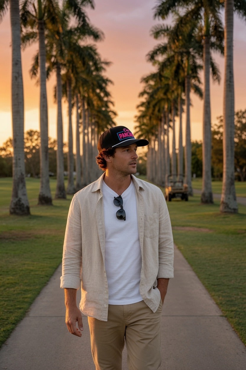 A man walking on a path lined with palm trees at sunset, wearing a beige overshirt over a white t-shirt, khaki pants, and a black 'DON PANCHITO NEON RECTANGLE BOX rope hat'.