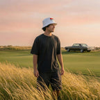 A man wearing the white DON PANCHITO SMOKE SHOW bucket hat, a black t-shirt, and black shorts stands in a field of tall golden grass on a golf course, with a vintage car in the background under a pinkish sunset sky.