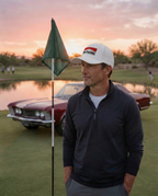 A man wearing the white DON PANCHITO SMOKE SHOW dad hat and a dark navy quarter-zip pullover stands on a golf course green near a pond, with a red classic car and a green flag in the background at sunset.