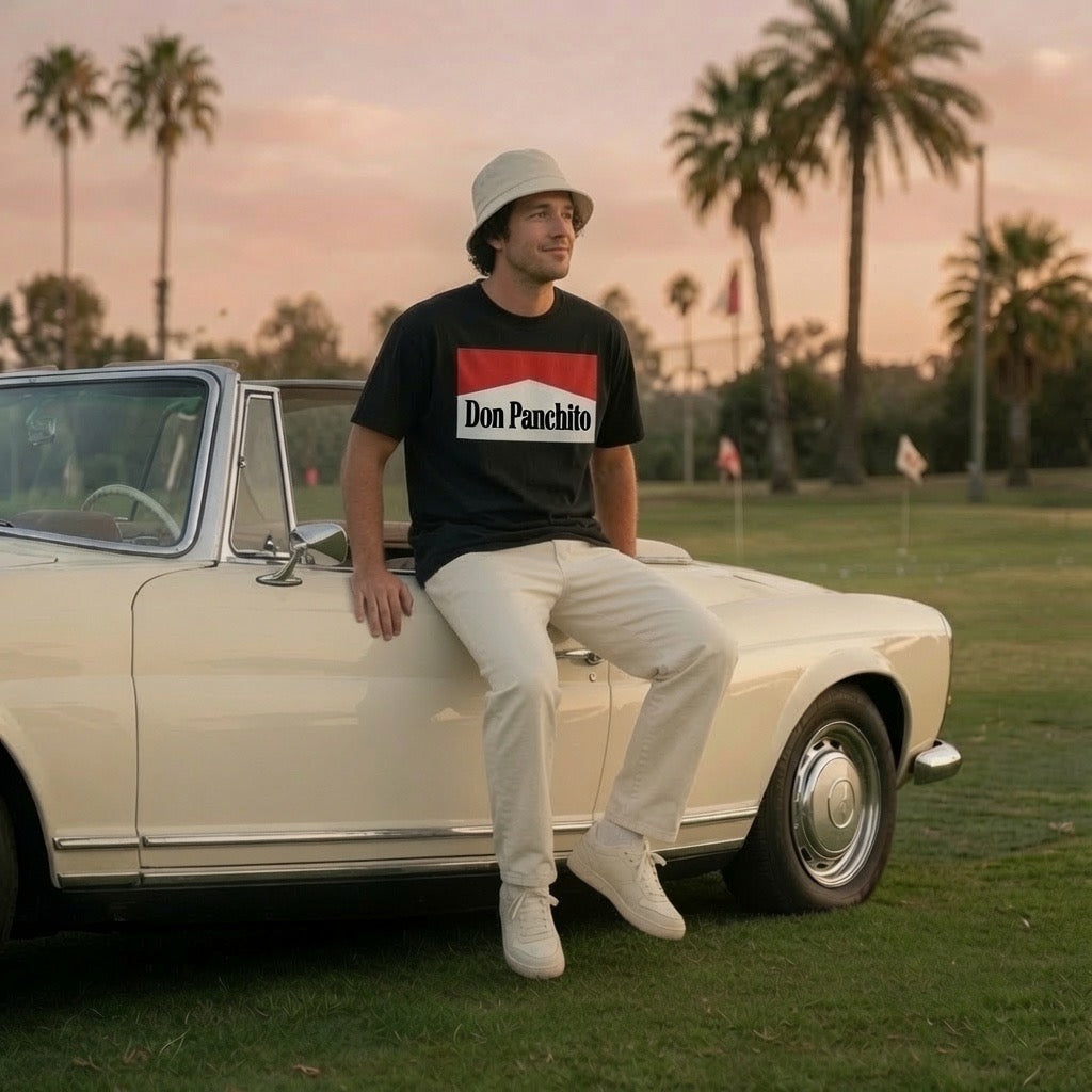 A man wearing the black DON PANCHITO SMOKE SHOW t-shirt, beige pants, and a bucket hat sits on the side of a cream-colored vintage convertible car on a grassy golf course at sunset.
