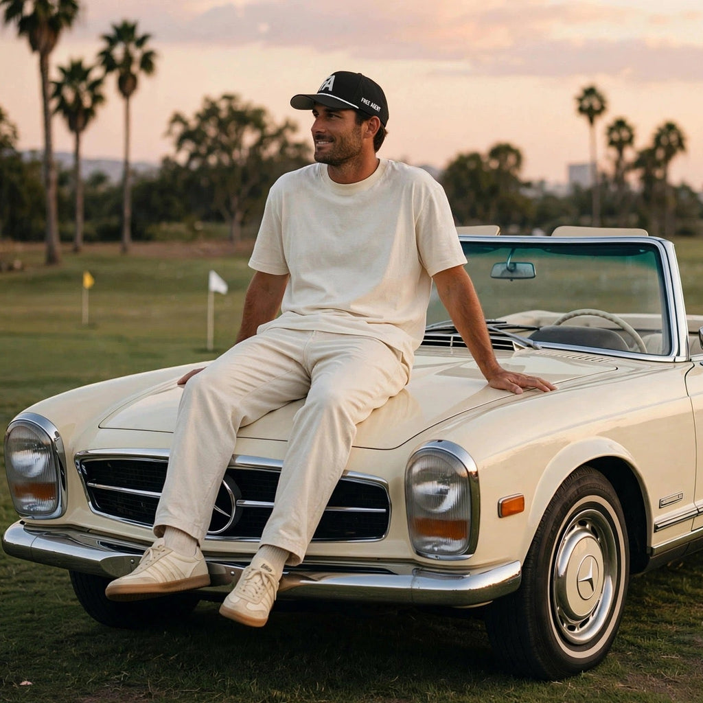 A man in a white t-shirt and matching pants sits on the hood of a cream convertible Mercedes-Benz car on a golf course at sunset, with a pyramid of golf balls on the grass beside it.