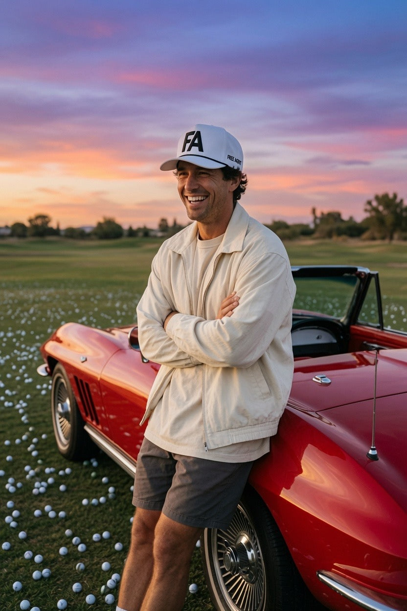 A smiling man wearing a white cap with a black 'FA' FREE AGENT logo, a cream jacket, and gray shorts leans against a red classic convertible car on a golf course covered in golf balls at sunset.
