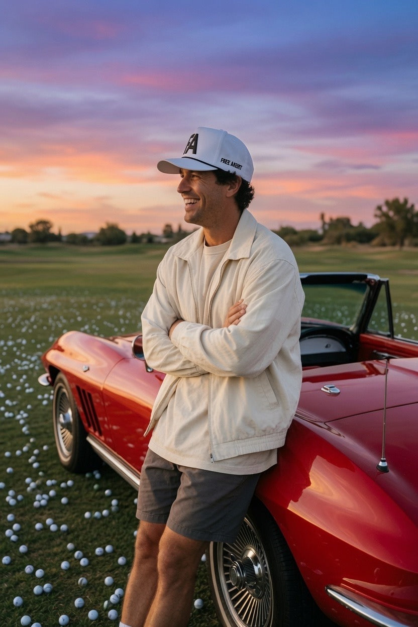 A man wearing a white "FREE AGENT FA" hat made by Don Panchito Golf, cream jacket, and gray shorts stands next to a red classic convertible car on a golf course at sunset, with golf balls scattered on the grass.