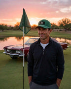 A man wearing the green FREE AGENT ALL BUSINESS rope hat and dark quarter-zip pullover stands on a golf course green with a flagpole in front of him and a classic car parked near a pond at sunset.
