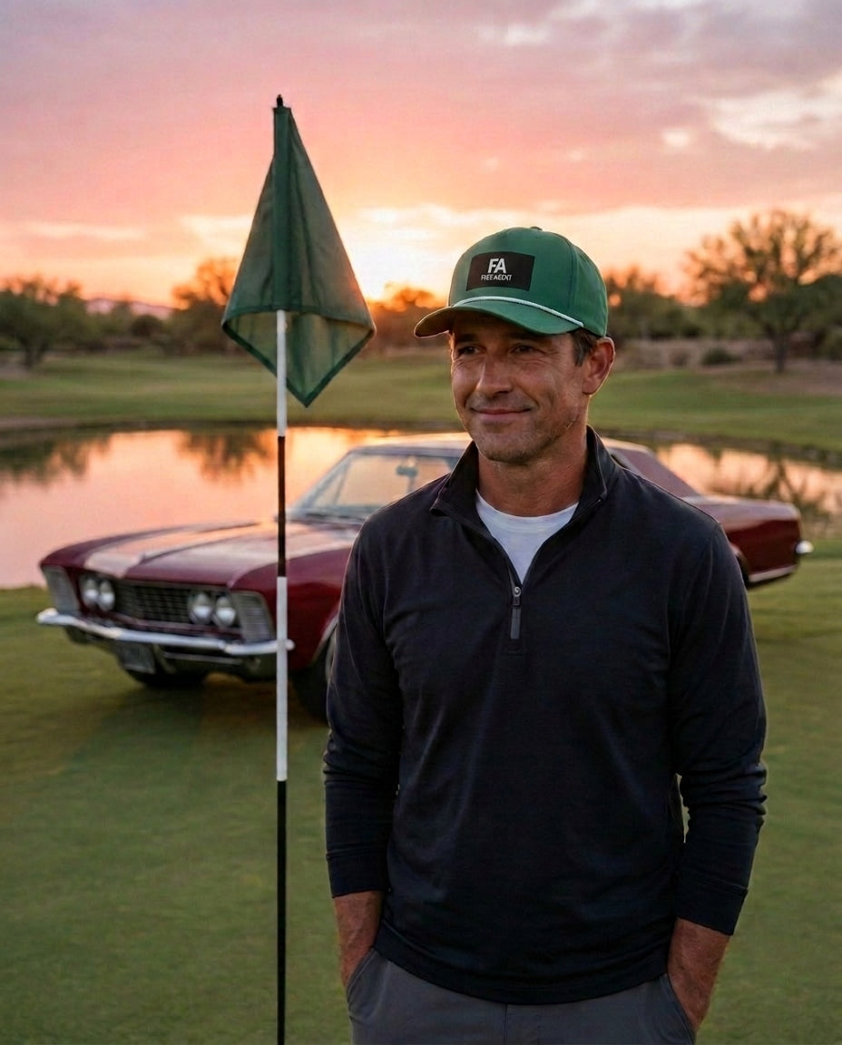 A man wearing the green FREE AGENT ALL BUSINESS rope hat and dark quarter-zip pullover stands on a golf course green with a flagpole in front of him and a classic car parked near a pond at sunset.