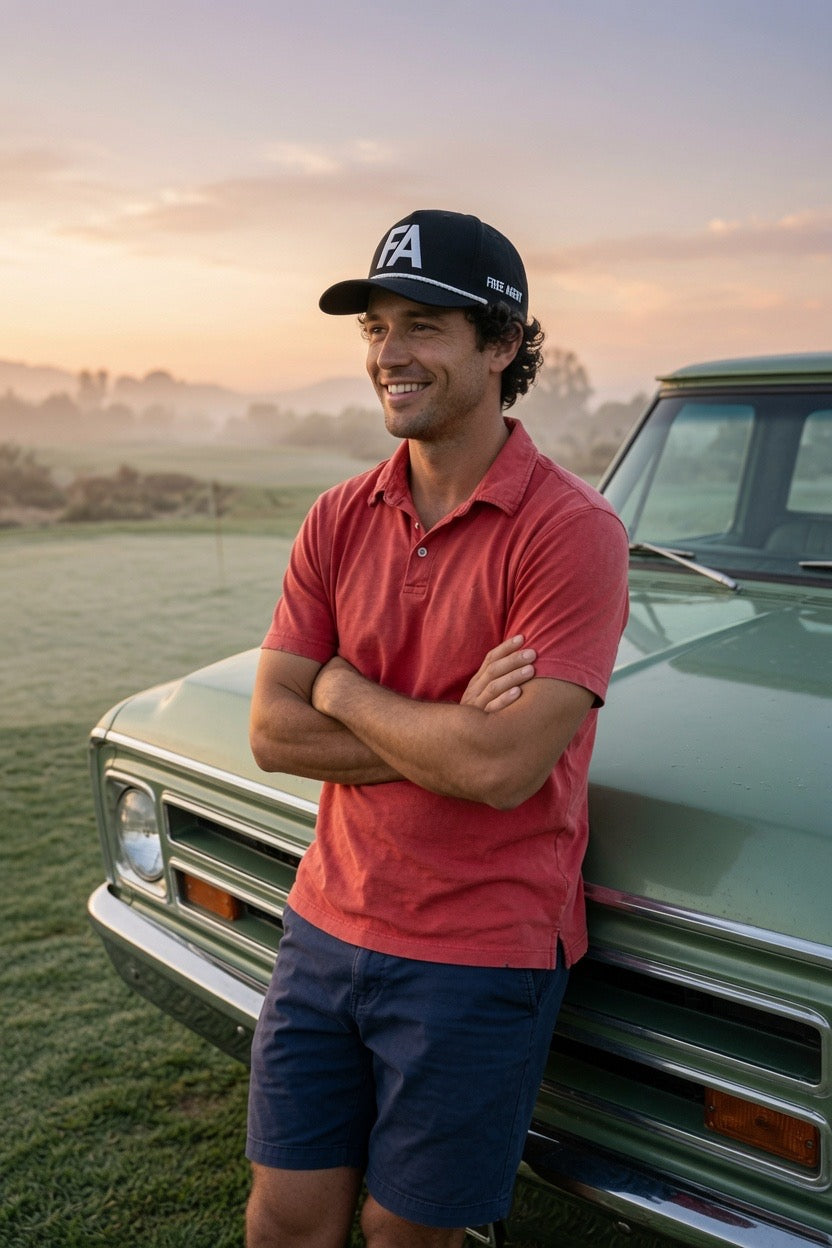 A man wearing a coral red polo shirt, navy shorts, and a black baseball cap with a white 'FA' FREE AGENT logo made by DON PANCHITO leans against the front of a vintage green truck in an outdoor field at sunset.