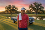 A man wearing a red polo shirt, navy pants, and the white FREE AGENT SCRIPT Rope Hat stands on a golf course at sunset with a blue pickup truck and water in the background.