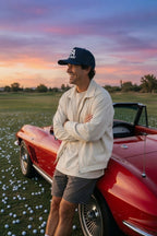 A man wearing a cream jacket, gray shorts, and a black baseball cap leans against a red convertible sports car on a grassy field scattered with golf balls under a colorful sunset sky.