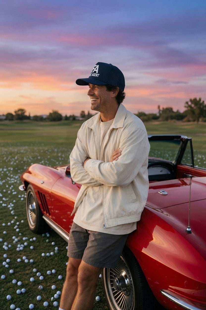 A man wearing a cream jacket, gray shorts, and a black baseball cap leans against a red convertible sports car on a grassy field scattered with golf balls under a colorful sunset sky.