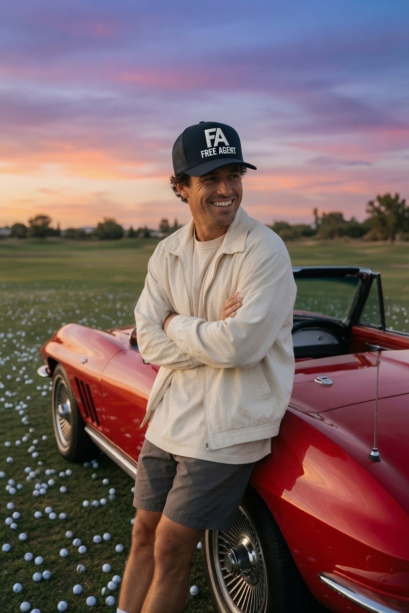A smiling man wearing a black 'FA FREE AGENT' cap, cream jacket, and gray shorts leans against a red classic convertible car on a golf course at sunset.