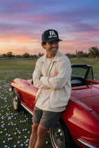 A smiling man wearing a black 'FA FREE AGENT' cap, cream jacket, and gray shorts leans against a red classic convertible car on a golf course at sunset.