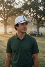 A man wearing a dark green polo shirt and the white GOLF NUT rope hat stands on a golf course at sunrise, with trees and a vintage car blurred in the background.