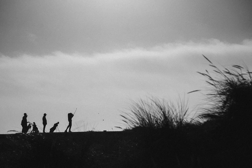 Black and white landscape photograph showing the silhouettes of several people standing on a grassy ridge against a cloudy sky, with tall grasses in the foreground.