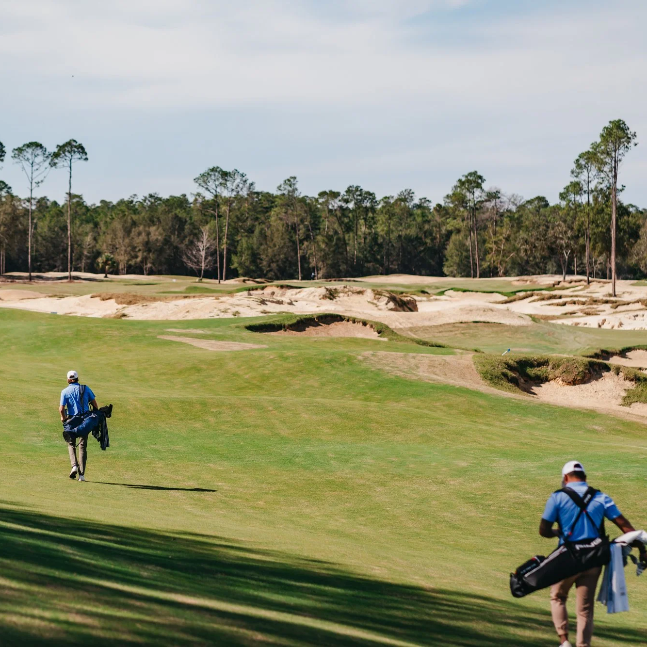 Two golfers walking across a grassy golf course with sand bunkers and trees in the background under a partly cloudy sky.